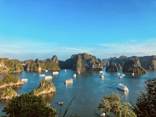 A scenic view of a bay with numerous limestone islands covered in lush greenery, surrounded by calm blue waters. Several boats and cruise ships are scattered across the water, indicating a popular tourist destination. The clear sky and bright sunlight enhance the vibrancy of the landscape.