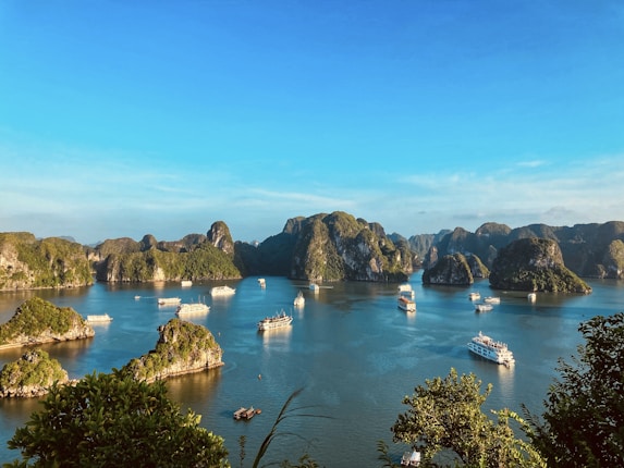 A scenic view of a bay with numerous limestone islands covered in lush greenery, surrounded by calm blue waters. Several boats and cruise ships are scattered across the water, indicating a popular tourist destination. The clear sky and bright sunlight enhance the vibrancy of the landscape.