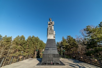 A large stone monument stands prominently against a clear blue sky, surrounded by dense green pine trees. The monument appears to be a statue of a person atop a tall, wide pedestal with inscriptions, and is placed on a raised platform with railings.