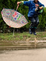 A child enthusiastically jumps into a muddy puddle, splashing water around. Wearing a blue jacket and rubber boots, the child is holding a colorful umbrella featuring animated characters. The background includes lush green foliage.