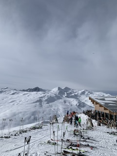 A snowy mountainous landscape with various skiers gathered around a ski lodge. Numerous ski poles and skis are scattered around, with the lodge positioned to the right of the image. The sky is overcast, and the surrounding mountains are covered in snow.