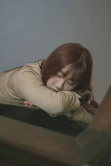 A person with long hair rests their head and arms on a surface, appearing to be in a deep sleep or contemplation. The lighting is soft and the setting seems calm and serene.