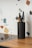 A black cylindrical knife holder containing several assorted knives is placed on a wooden kitchen countertop. The background features a terrazzo-style wall pattern with colorful fragments in shades of blue, orange, and black.