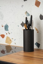A black cylindrical knife holder containing several assorted knives is placed on a wooden kitchen countertop. The background features a terrazzo-style wall pattern with colorful fragments in shades of blue, orange, and black.