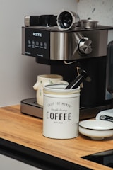 A modern espresso machine with various controls and features is positioned on a wooden countertop. Nearby, a white container labeled 'Enjoy the Moment, Simple Living COFFEE' holds a black coffee scoop. A ceramic mug with a floral pattern is beside the machine, and a small white dish is also present.
