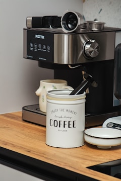 A modern espresso machine with various controls and features is positioned on a wooden countertop. Nearby, a white container labeled 'Enjoy the Moment, Simple Living COFFEE' holds a black coffee scoop. A ceramic mug with a floral pattern is beside the machine, and a small white dish is also present.