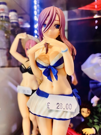 A colorful anime figure is posed with hands near the face, wearing a blue and white sailor-themed outfit. A price tag displays '£28.00'. There are other anime figures in the background, positioned on shelves with various lighting effects.