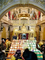A richly decorated temple interior with an elaborate altar, adorned with gold and intricate sculptures. Numerous trays filled with a variety of sweets and offerings are prominently displayed in the foreground. The ambiance is vibrant with people seen moving around, participating in what appears to be a cultural or religious event.