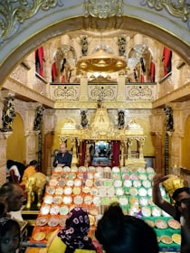 A richly decorated temple interior with an elaborate altar, adorned with gold and intricate sculptures. Numerous trays filled with a variety of sweets and offerings are prominently displayed in the foreground. The ambiance is vibrant with people seen moving around, participating in what appears to be a cultural or religious event.