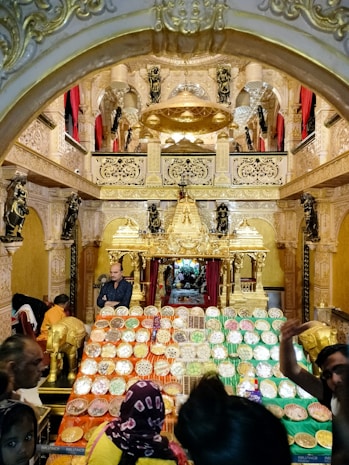 A richly decorated temple interior with an elaborate altar, adorned with gold and intricate sculptures. Numerous trays filled with a variety of sweets and offerings are prominently displayed in the foreground. The ambiance is vibrant with people seen moving around, participating in what appears to be a cultural or religious event.