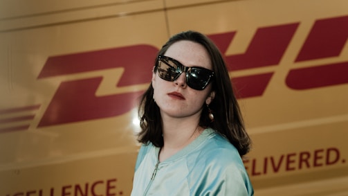 A woman wearing large sunglasses is standing in front of a yellow van with a prominent red logo. She has shoulder-length brown hair and is wearing pearl earrings. The sunlight casts a shadow, giving the scene a dramatic flair.