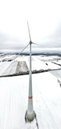 A tall wind turbine stands in the middle of a vast, snow-covered landscape, with fields extending out to the horizon. The sky appears overcast, creating a muted and serene atmosphere.