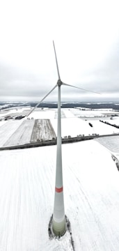A tall wind turbine stands in the middle of a vast, snow-covered landscape, with fields extending out to the horizon. The sky appears overcast, creating a muted and serene atmosphere.