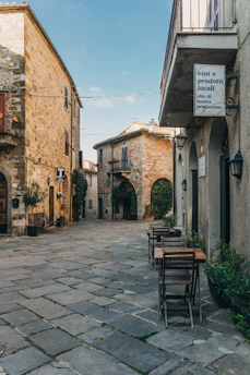 A picturesque cobblestone street in a quaint village, lined with rustic stone buildings. The scene features outdoor seating with wooden tables and chairs against the walls of the buildings. A sign advertising local wine and products hangs above one of the doorways. The atmosphere is serene and timeless, with climbing plants adding to the charm of the architecture.