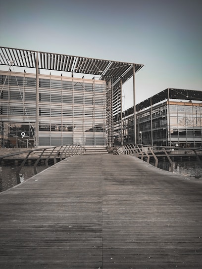 A modern architectural structure with glass façades and metal frameworks. A wooden walkway leads up to the building, crossing over a water feature. The building has a sleek, geometric design with a large overhanging roof.