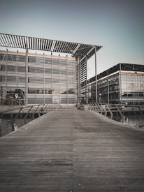 A modern architectural structure with glass façades and metal frameworks. A wooden walkway leads up to the building, crossing over a water feature. The building has a sleek, geometric design with a large overhanging roof.