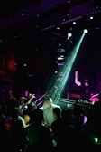 A DJ is performing on stage under bright lights, with a crowd gathered in front of the DJ booth. The atmosphere is lively, with people dancing and interacting energetically. The lighting creates a nightclub ambiance with colorful beams illuminating the scene.