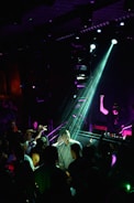 A DJ is performing on stage under bright lights, with a crowd gathered in front of the DJ booth. The atmosphere is lively, with people dancing and interacting energetically. The lighting creates a nightclub ambiance with colorful beams illuminating the scene.
