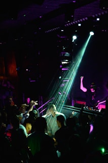 A DJ is performing on stage under bright lights, with a crowd gathered in front of the DJ booth. The atmosphere is lively, with people dancing and interacting energetically. The lighting creates a nightclub ambiance with colorful beams illuminating the scene.