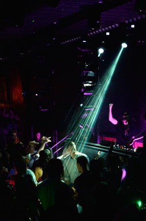 A DJ is performing on stage under bright lights, with a crowd gathered in front of the DJ booth. The atmosphere is lively, with people dancing and interacting energetically. The lighting creates a nightclub ambiance with colorful beams illuminating the scene.