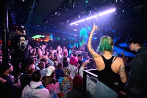 A crowded nightclub scene with people dancing and enjoying a live music performance. Neon and blue lights illuminate the room, creating an energetic atmosphere. A woman with green hair and tattoos raises her arm in excitement amidst the audience.