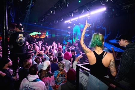A crowded nightclub scene with people dancing and enjoying a live music performance. Neon and blue lights illuminate the room, creating an energetic atmosphere. A woman with green hair and tattoos raises her arm in excitement amidst the audience.