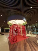 A large decorative red structure adorned with lights stands in the center of a spacious, modern indoor area. The structure resembles a wrapped gift box with a heart design on its side, surrounded by warm ambient lighting and wooden flooring. Overhead, there is an illuminated circular ceiling with greenery.