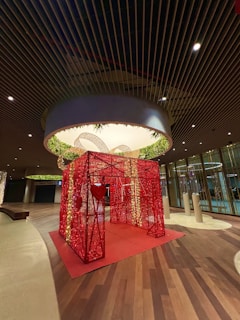 A large decorative red structure adorned with lights stands in the center of a spacious, modern indoor area. The structure resembles a wrapped gift box with a heart design on its side, surrounded by warm ambient lighting and wooden flooring. Overhead, there is an illuminated circular ceiling with greenery.