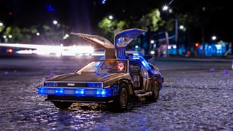 A toy version of a DeLorean car with its gull-wing doors open, illuminated by blue lights. The car is placed on a textured gray surface, likely a street, with a background featuring blurred city lights and greenery.