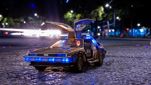 A toy version of a DeLorean car with its gull-wing doors open, illuminated by blue lights. The car is placed on a textured gray surface, likely a street, with a background featuring blurred city lights and greenery.
