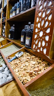 Wooden shelves display large jars containing various spices. In the foreground, open compartments hold an assortment of spices and ingredients, such as dried herbs, large stones, and a pile of coarse orange-brown chunks. A blue-handled metal scoop is resting in one of the compartments. The carved latticework on the side adds an ornamental touch.