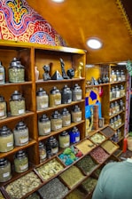 A colorful spice shop interior features wooden shelves lined with numerous glass jars containing various spices and herbs. The jars are organized neatly, with some open trays displaying dried leaves and colorful grains. Above the shelves are intricately painted, vibrant patterns in red, blue, and yellow. Small statues and decorative items are displayed amid the jars, adding to the eclectic aesthetic.