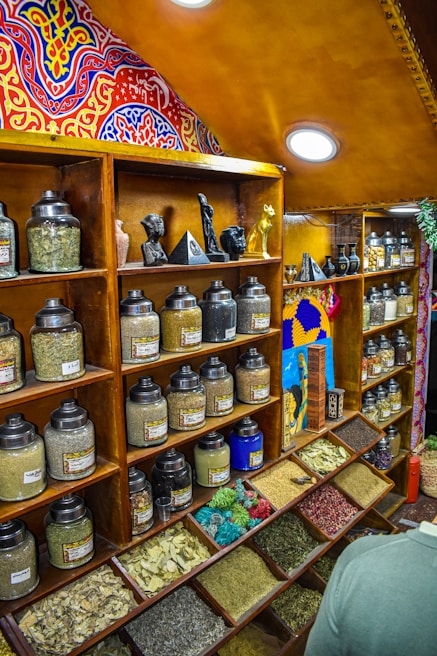 A colorful spice shop interior features wooden shelves lined with numerous glass jars containing various spices and herbs. The jars are organized neatly, with some open trays displaying dried leaves and colorful grains. Above the shelves are intricately painted, vibrant patterns in red, blue, and yellow. Small statues and decorative items are displayed amid the jars, adding to the eclectic aesthetic.