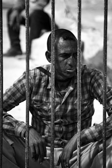 A man sits behind metal bars, wearing a plaid shirt. His expression appears somber, suggesting a contemplative or resigned mood. The image is in black and white, highlighting the contrast and texture of the scene.