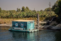A floating pump station is situated on a body of water, surrounded by lush greenery and banana trees. The structure is painted turquoise and white, with flags displayed on its side. Large industrial pipes extend from the station, suggesting its role in water management or irrigation. The calm water reflects parts of the station, creating a serene atmosphere, while the clear sky above provides a sense of openness.