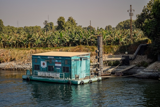A floating pump station is situated on a body of water, surrounded by lush greenery and banana trees. The structure is painted turquoise and white, with flags displayed on its side. Large industrial pipes extend from the station, suggesting its role in water management or irrigation. The calm water reflects parts of the station, creating a serene atmosphere, while the clear sky above provides a sense of openness.