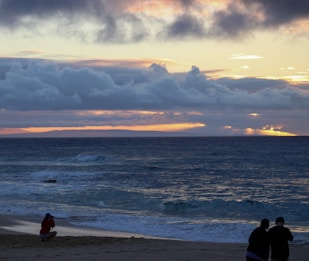 A scenic beach view with a vast ocean stretching toward the horizon under a dramatic sky. The sky is filled with dense, dark clouds tinged with orange and pink hues from the setting sun. On the sandy shore, one person is crouching and appears to be photographing the scene, while two others are standing and possibly engaging in conversation or viewing something together.