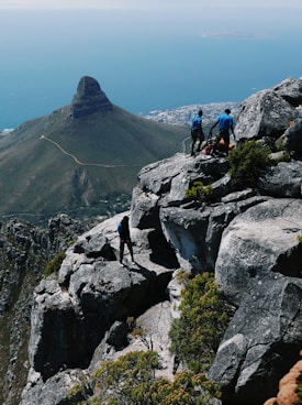 A group of climbers stands on rocky terrain overlooking a vast ocean and a distinctive, steep-sided mountain. The landscape includes lush greenery and a clear view of a distant city, suggesting a high-altitude vantage point.