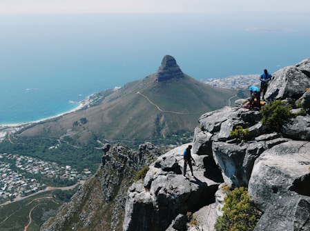 A dramatic landscape with a rugged mountain ridge in the foreground where several people are engaged in climbing activities. The background features a distinct conical hill with cityscape elements at its base and a vast ocean stretching to the horizon.