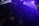 A dimly lit nightclub or dance venue filled with people. Colored lights and beams cast a blue and purple ambiance over the crowd. Some people are dancing, and others are holding drinks. The atmosphere is lively and energetic.