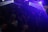 A dimly lit nightclub or dance venue filled with people. Colored lights and beams cast a blue and purple ambiance over the crowd. Some people are dancing, and others are holding drinks. The atmosphere is lively and energetic.