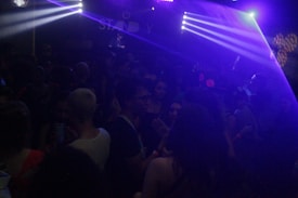 A dimly lit nightclub or dance venue filled with people. Colored lights and beams cast a blue and purple ambiance over the crowd. Some people are dancing, and others are holding drinks. The atmosphere is lively and energetic.