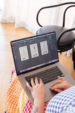 A person is using a laptop, working on graphic design, with three similar-looking design templates on the screen. The setting appears to be indoors with natural light coming through sheer curtains. The person has brightly painted nails and a tattooed forearm, wearing a colorful checked fabric.