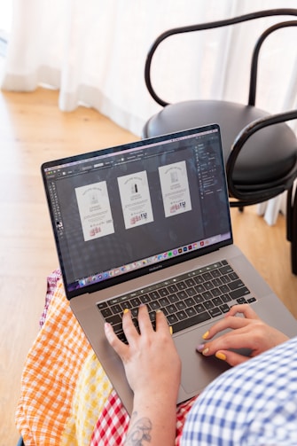 A person is using a laptop, working on graphic design, with three similar-looking design templates on the screen. The setting appears to be indoors with natural light coming through sheer curtains. The person has brightly painted nails and a tattooed forearm, wearing a colorful checked fabric.