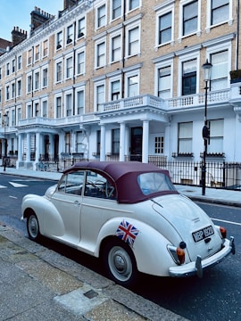 A vintage white car with a maroon soft top is parked on the street in front of a row of classic, multi-storey townhouses. The car has a Union Jack sticker on the rear and features a retro design, complementing the traditional architecture of the buildings behind.