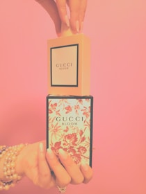 A pair of hands elegantly display two Gucci Bloom perfume boxes, one atop the other against a soft pink background. The lower box features a floral design with red blossoms on a light blue background, while the upper box is plain with a minimalist aesthetic. Both hands are adorned with pearl bracelets and rings, and have neatly manicured nails.