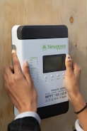 A person is installing or interacting with a solar charge controller mounted on a wooden surface. The device is labeled 'Newpowa' with various technical specifications visible, including 'MPPT 100 | 40', and it features a small screen and several buttons.