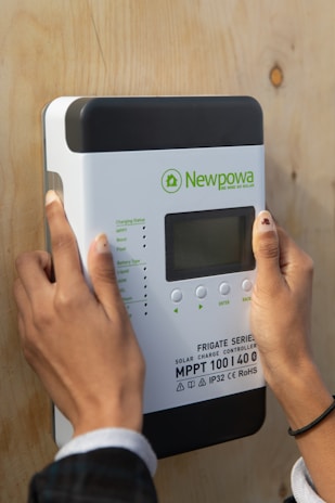 A person is installing or interacting with a solar charge controller mounted on a wooden surface. The device is labeled 'Newpowa' with various technical specifications visible, including 'MPPT 100 | 40', and it features a small screen and several buttons.