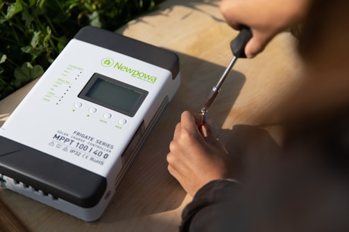 A person is using a screwdriver to work on a solar charge controller device labeled 'Newpowa.' The device is placed on a wooden surface, and greenery is visible in the background.
