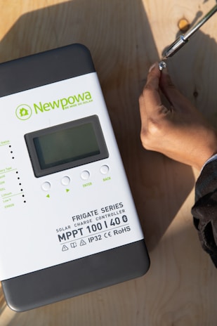 A solar charge controller from the Newpowa brand labeled as the Frigate Series MPPT 100 | 40 is displayed on a wooden surface. A hand is shown using a screwdriver to adjust or tighten something on the surface.